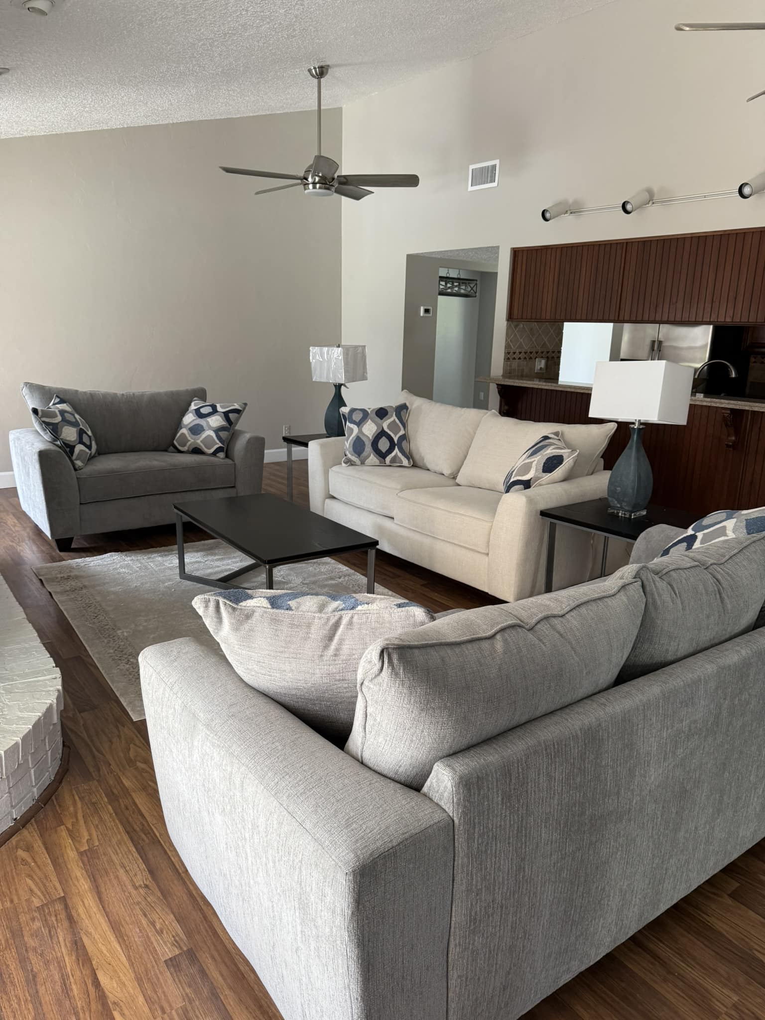 Modern living room with two gray sofas and one cream sofa, accented by blue patterned cushions. Wooden floors, ceiling fan, and neutral walls. Cozy, sleek ambiance.
