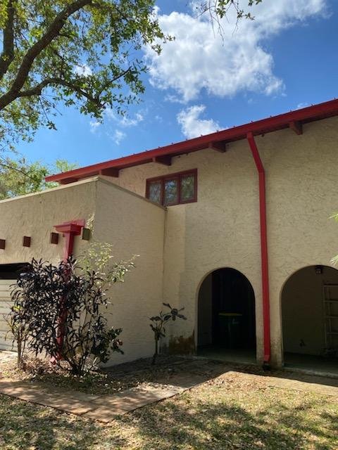 A beige two-story house with red trim features arched doorways and a garage. A leafy tree frames the top, with a vibrant blue sky and cloud above.