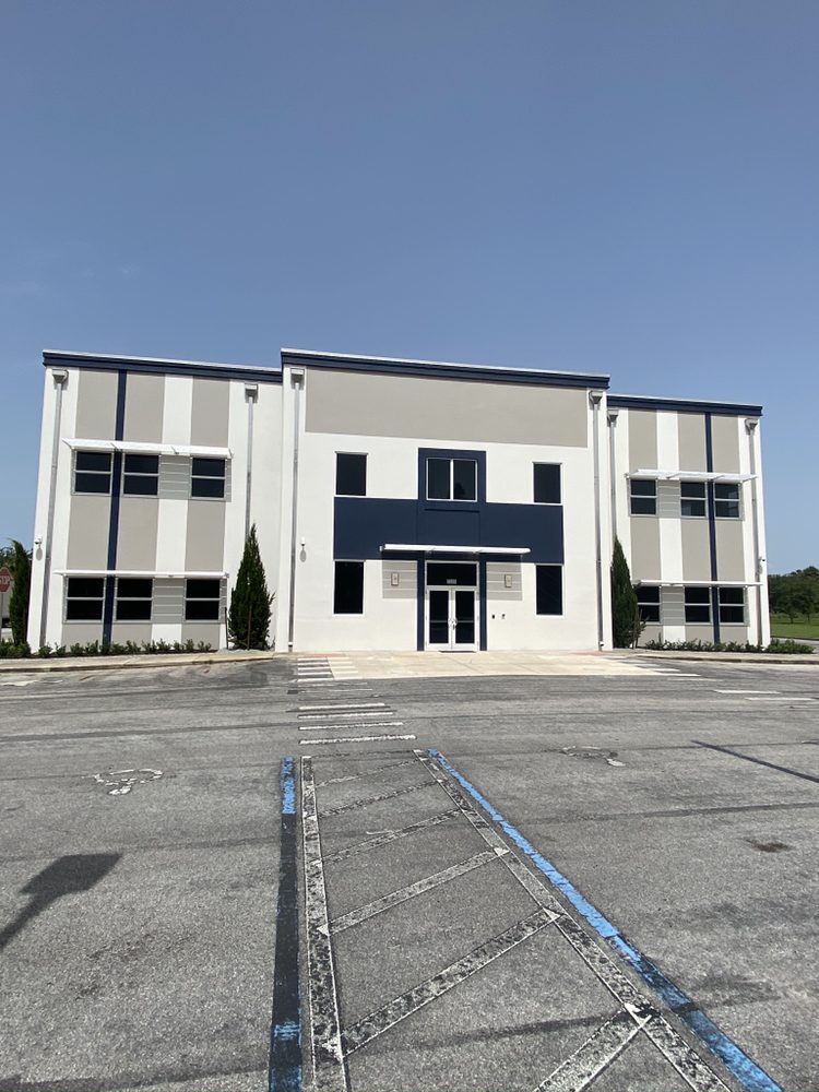 Modern two-story building with a symmetrical facade, featuring large windows and a minimalistic design. Empty parking lot in the foreground under clear skies.