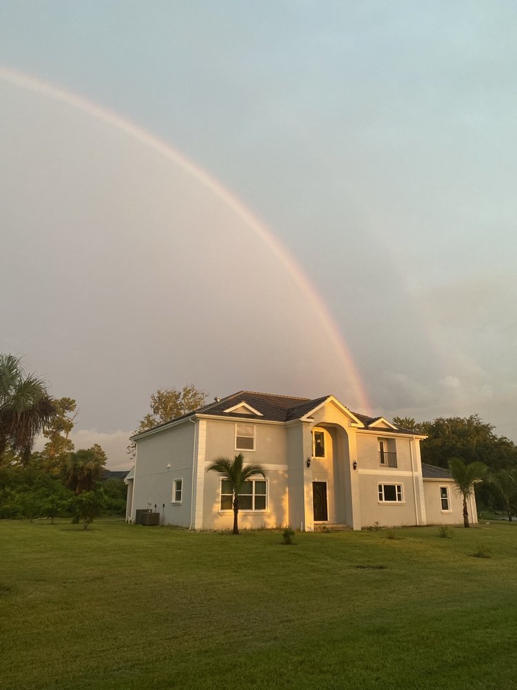 A serene image of a two-story white house on a lush green lawn, illuminated by a warm glow. A faint rainbow arcs gracefully over the clear sky.