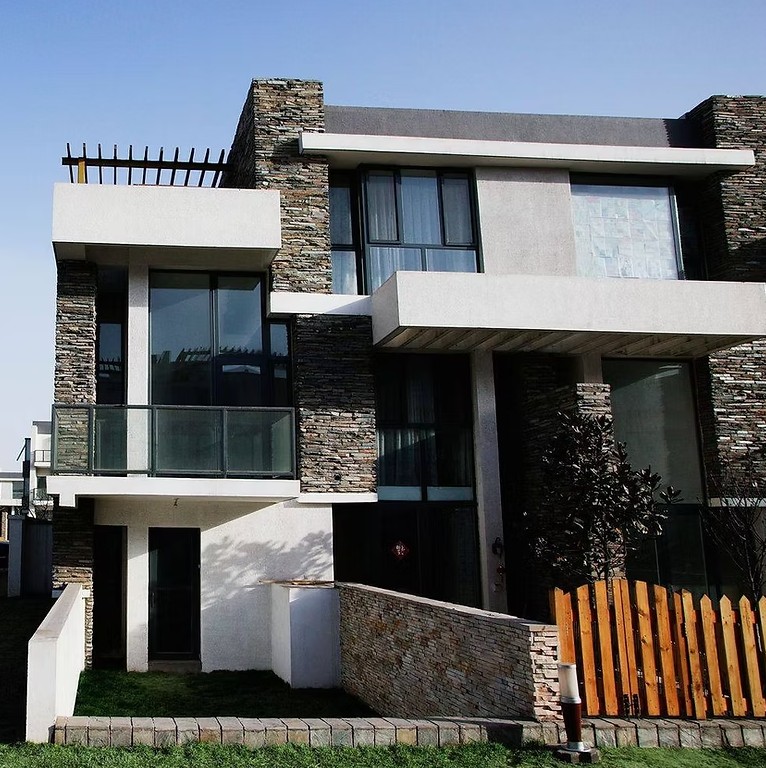 Modern two-story house with stone and stucco facade, large glass windows, a wooden fence, small lawn, and blue sky, conveying a sleek, contemporary vibe.